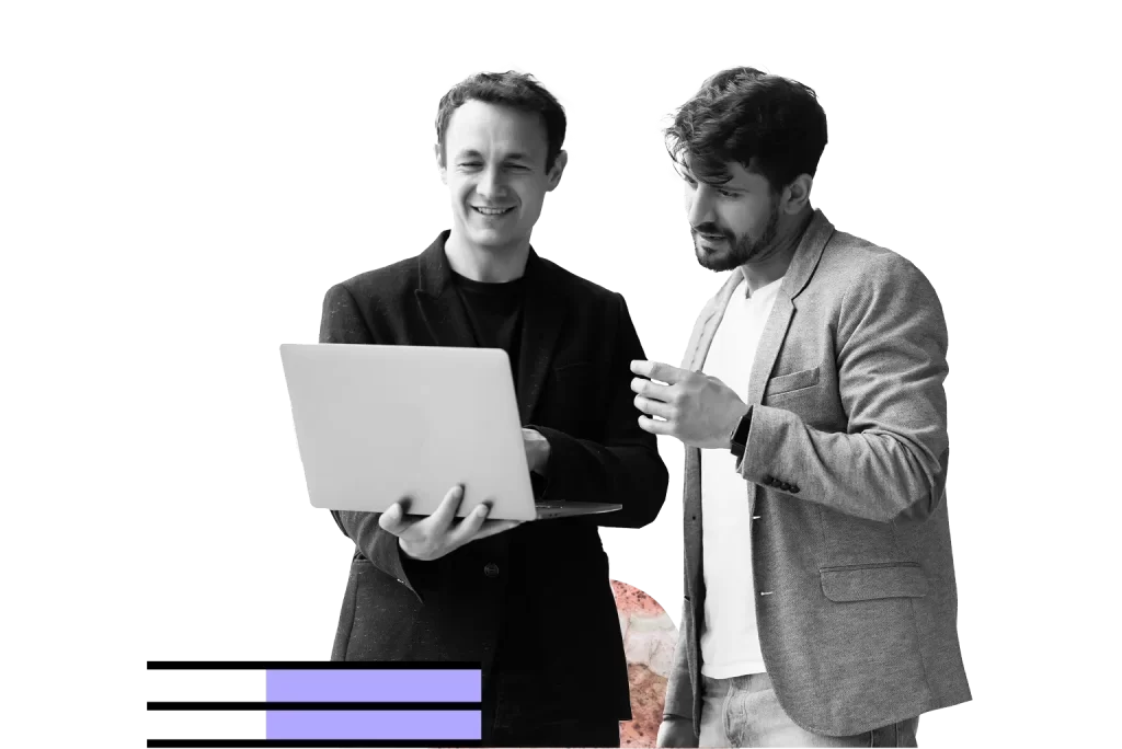 Two men standing and looking at a laptop, one of them holding a cup. Both are wearing blazers and smiling, engaged in conversation.