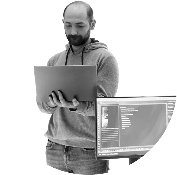 A man in a hoodie holds a laptop and looks at the screen; a computer monitor displaying code is visible beside him.